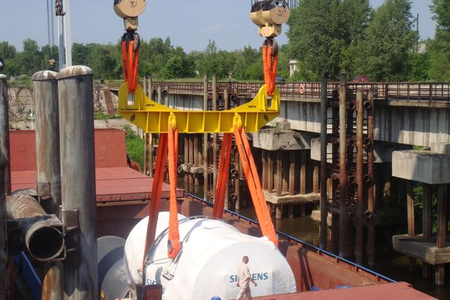 A crane has two synthetic orange lifting slings attached to it. The slings are attached to lifting points on a large metal vessel being pulled from a ship.