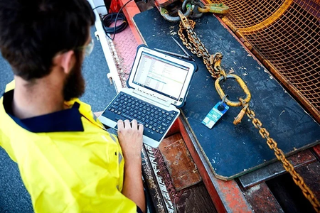 A man in a CERTEX Lifting uniform is using the Motion Kinetic Asset Management System on a computer to conduct a thorough safety inspection of a lifting and rigging chain. 