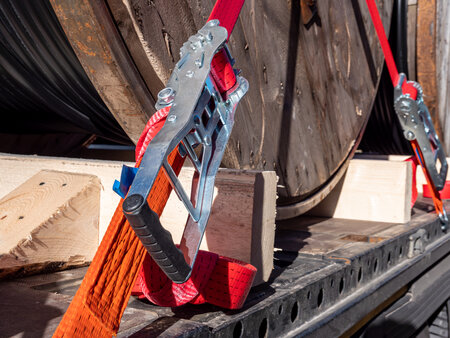A close-up image of a webbing ratchet tied down, securing a load of steel rope spools on the back of a semi-trailer.